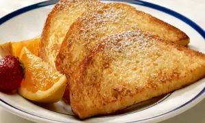 Fattiga riddare, gyllene och pudrade med florsocker, serveras med färska jordgubbar och apelsinklyftor på en vit tallrik med blå kant.