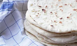 En hög med nybakade tortillabröd, perfekta för ditt nästa tortilla recept. Bredvid ligger en blåvitrutig kökshandduk.