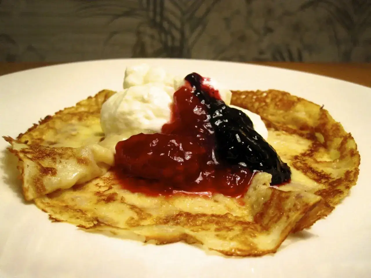 En gyllene pannkaka toppad med vispad grädde, hallonsylt och blåbärssylt. Ett perfekt pannkakor recept för en mysig fika.