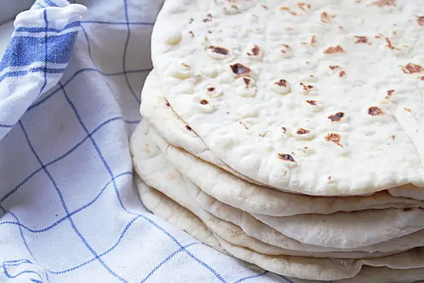 En hög med nybakade tortillabröd, perfekta för ditt nästa tortilla recept. Bredvid ligger en blåvitrutig kökshandduk.